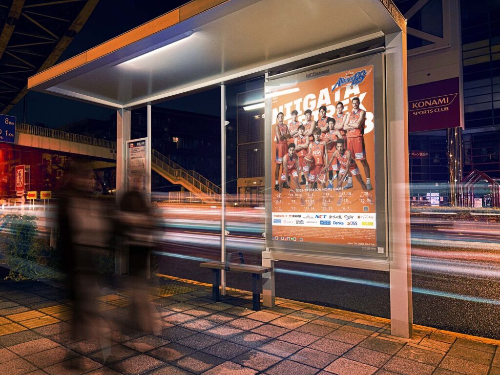 夜のバス停に掲出されたバスケットボールチームのポスター。チームが一体となったビジュアルが、光の軌跡とともに都市の活気を演出している