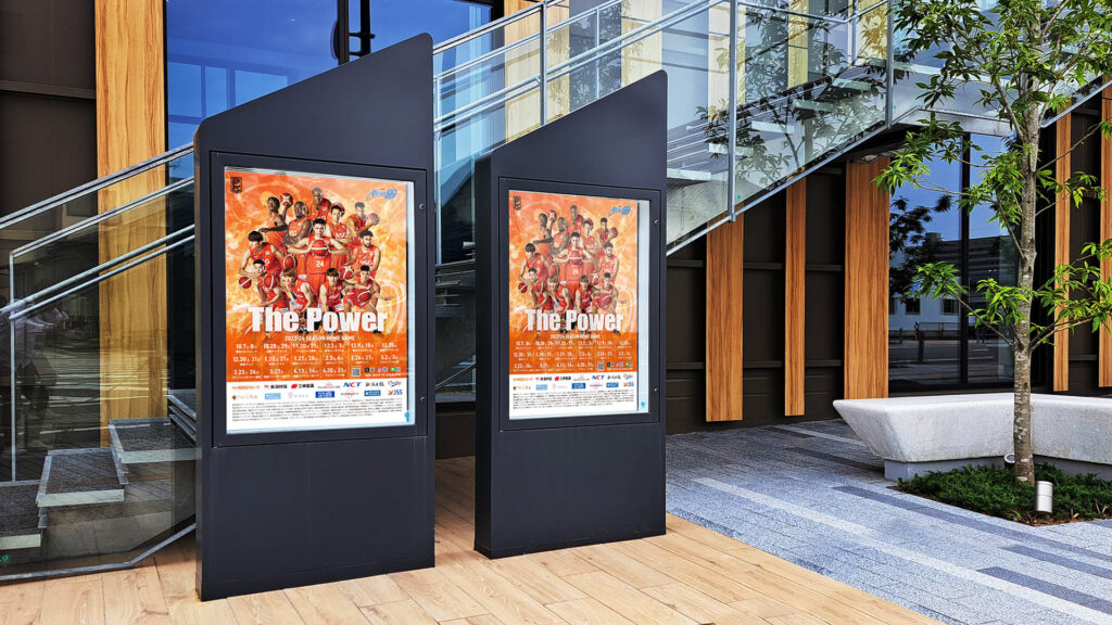 現代的な建物の前に設置されたデジタルサイネージに、バスケットボールチームのポスターが2面掲出されている。オレンジを基調としたデザインが力強さと一体感を表現している